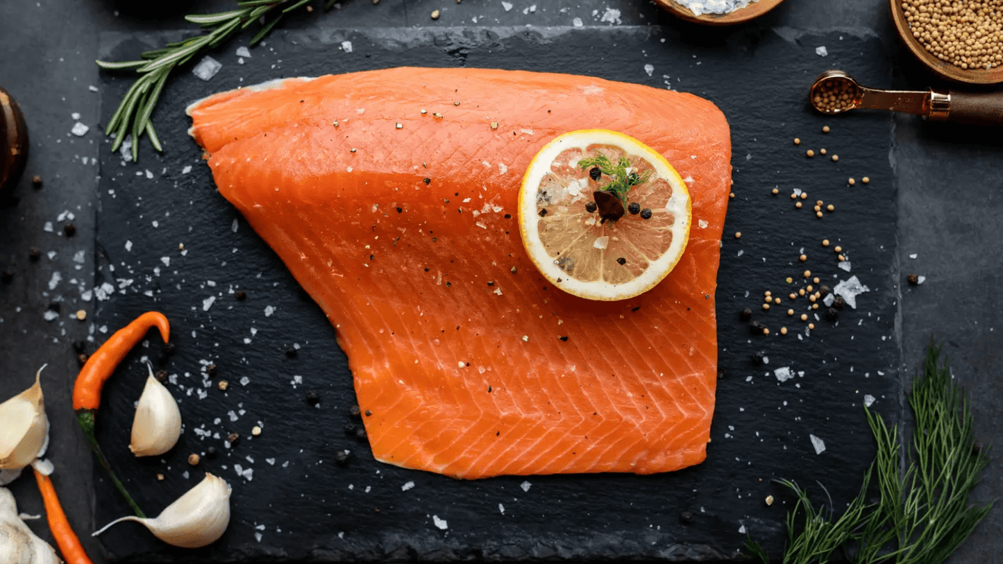 Fresh salmon fillet with lemon, garlic, and chili on a slate background, garnished with herbs and spices.
