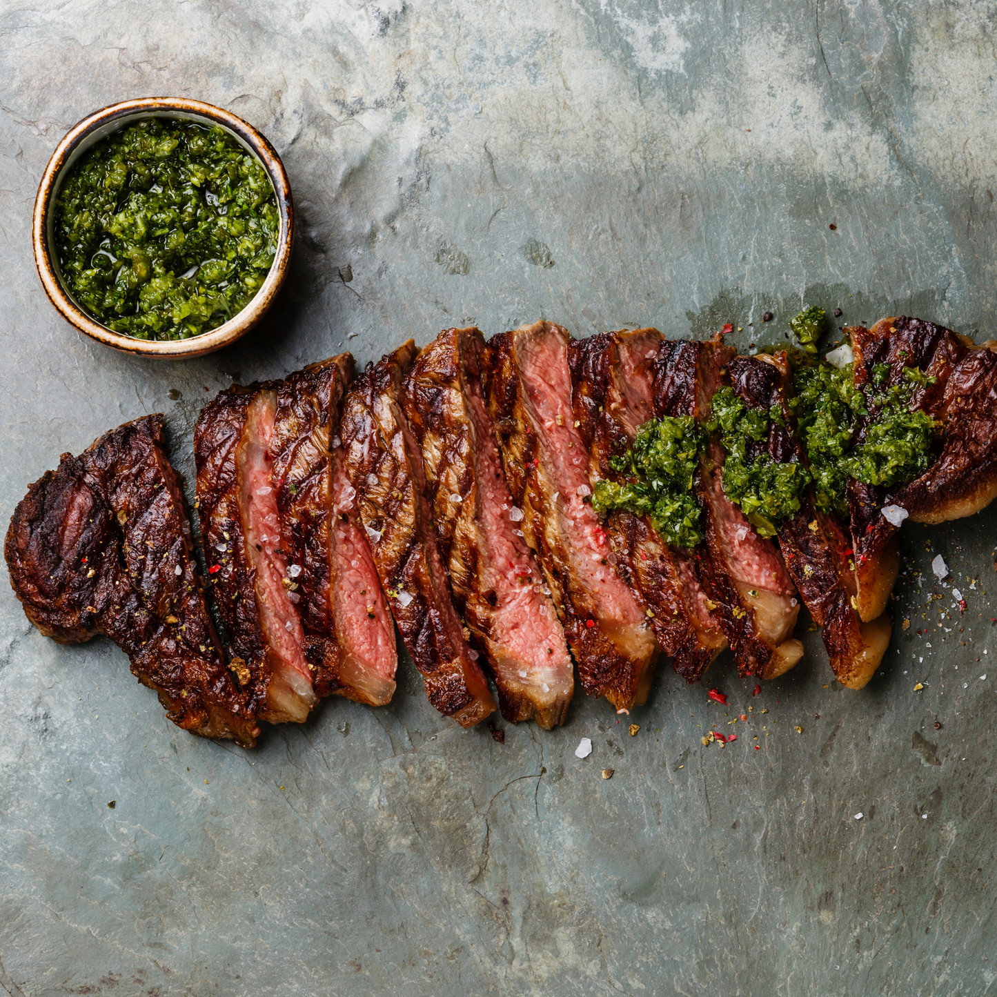 Sliced grass-fed beef steak topped with chimichurri sauce, served with a side of green sauce.
