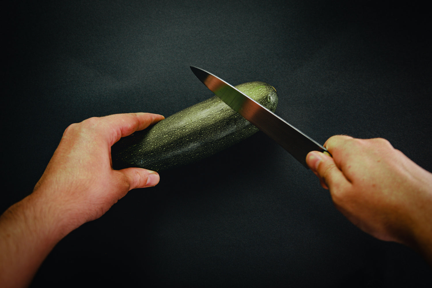 Person slicing a fresh zucchini with a sharp knife on a dark background.