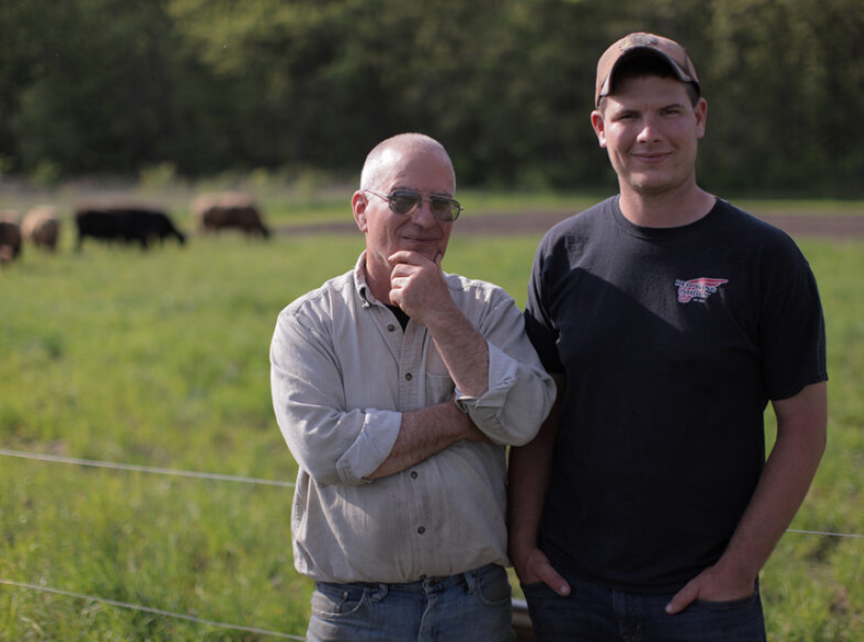 Two farmers posing together in a green pasture with cattle in the background, showcasing rural life and farming.